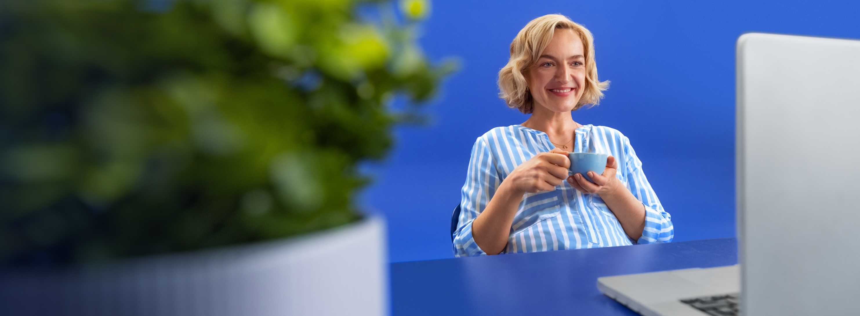 Une femme aux cheveux blonds et à la chemise rayée bleu et blanc est assise avec une tasse de café devant un ordinateur portable, sur un fond bleu vif.