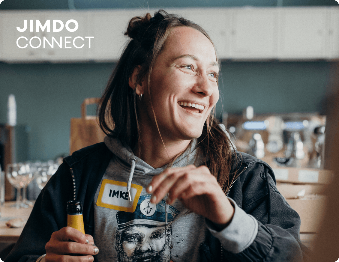 A smiling woman with long blonde hair, sitting at a table with a name badge and a drink in her hand, representing networking at a JIMDOConnect event.